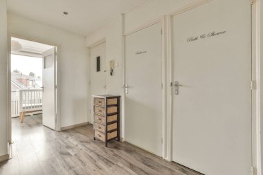 Beyaz duvarları ve kapıları olan koridor, modern dairede pencereleri ve parke zemini olan geniş bir odaya açılıyor.