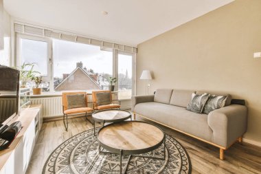 Televizyonun önündeki gri kanepe ve pencerenin karşısındaki yemek masası ile aydınlık oturma odasının basit bir iç mekanı.