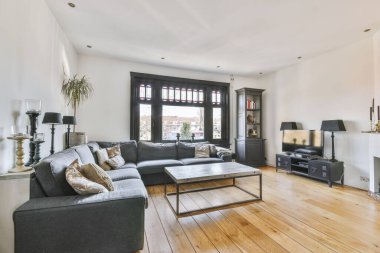 Geniş siyah çerçeveli pencereleri ve TV karşısında gri koltuğu olan basit bir oturma odası.