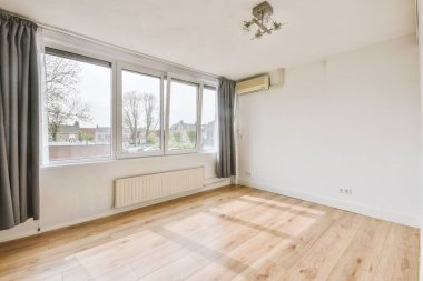 Pencereleri, gri perdeleri, radyatörü, avizesi ve kliması olan ferah, boş bir oda.