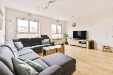 Televizyonun önündeki gri kanepe ve pencerenin karşısındaki yemek masası ile aydınlık oturma odasının basit bir iç mekanı.