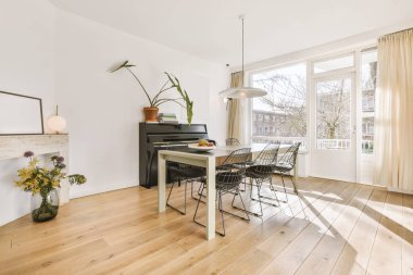 Modern odanın içi pencerelerden ve kapılardan gelen güneş ışığıyla aydınlanıyor. Duvara dayanmış bir piyanoyla minimalist bir yemek odasında.