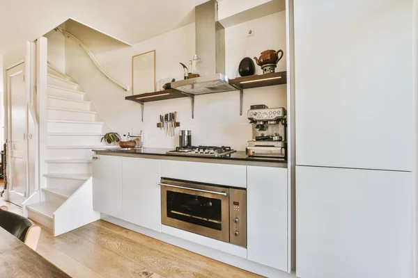 A fragment of the interior of a light home kitchen in a minimalist style with a sink, stove, extractor hood and with white furniture next to the stairs