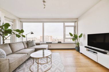The interior of a modern living room with large panoramic windows, a comfortable sofa, a stylish coffee table and a TV on a pedestal against the wall opposite