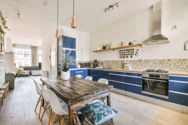 Interior of contemporary kitchen with stylish blue cupboards and appliances near dining area in contemporary apartment