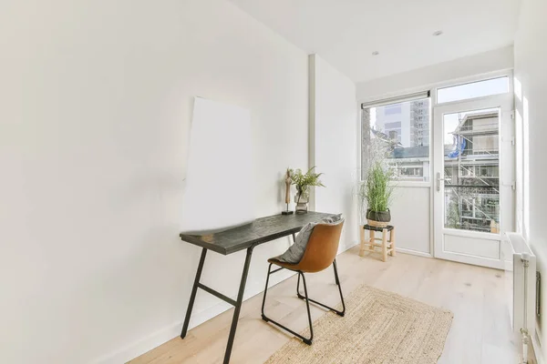 Narrow bright room with a balcony door, a table and a chair against a white wall