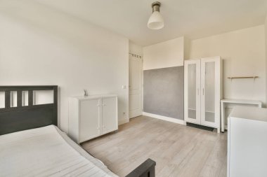 Küçük yatak odasının minimalist iç tasarımı. Köşesinde yumuşak yatak, ahşap gardırop ve lamba aydınlatmalı raflar var.