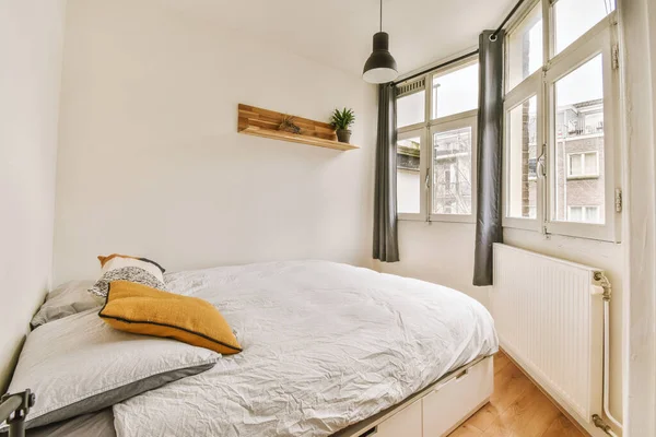 Minimalist interior design of small bedroom with soft bed placed in corner and wooden wardrobe and shelves with lamp illumination