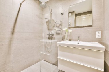 Modern banyoda aynalı lavabo ve cam kapılı duş kutusu ve beyaz fayanslı duvarlar.