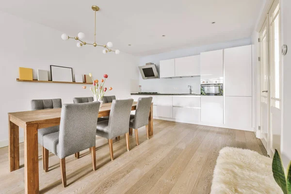 Modern minimalist style interior design of studio apartment with open white kitchen and dining zone with table and chairs illuminated by loft style lamp