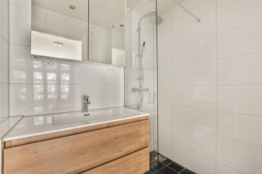 Modern banyoda aynalı lavabo ve cam kapılı duş kutusu ve beyaz fayanslı duvarlar.