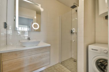 Aynalı lavabo ve çamaşır makinesi modern banyodaki cam kapılı duş kutusunun yanında beyaz fayanslı duvarları var.