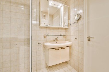 Modern banyoda aynalı lavabo ve cam kapılı duş kutusu ve beyaz fayanslı duvarlar.