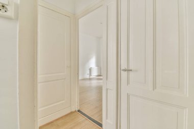 Beyaz duvarları ve kapıları olan dar koridor modern dairede pencereleri ve parke zemini olan geniş odaya açılıyor.