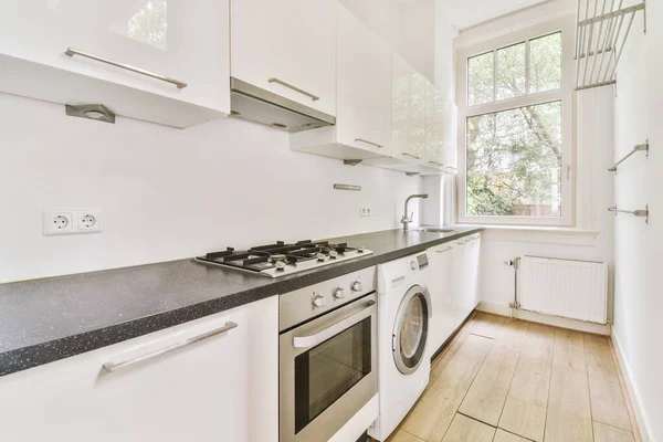 Well organized small home kitchen interior with sink under window and light furniture with stove and fridge in urban apartment