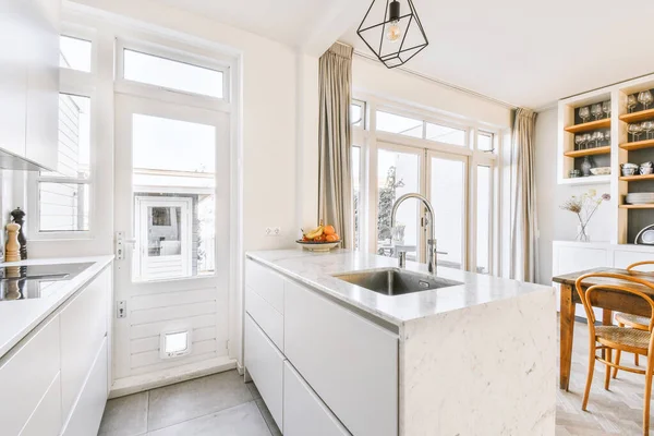 Interior of spacious light dining room with elegant chandelier located near kitchen with comfortable furniture and appliances