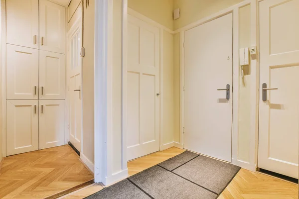 Narrow corridor with white walls and doors leading to spacious room with windows and parquet floor in modern apartment