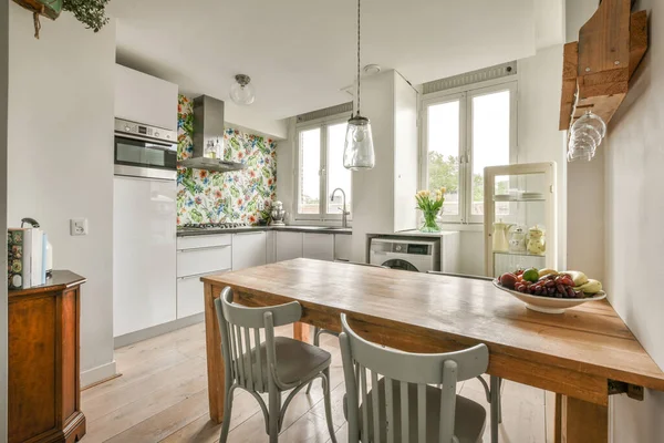 Modern minimalist style interior design of studio apartment with open white kitchen and dining zone with table and chairs illuminated by loft style lamp