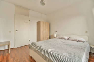 Beyaz çarşaflı rahat bir yatak ve lambasıyla pencere ve balkon kapısının yakınına yerleştirilmiş aydınlık yatak odası.