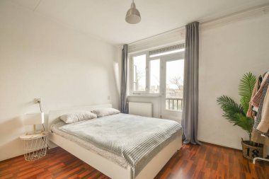 Beyaz çarşaflı rahat bir yatak ve lambasıyla pencere ve balkon kapısının yakınına yerleştirilmiş aydınlık yatak odası.