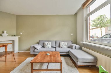 Büyük pencerenin yanında halısı ve bej koltuğu olan basit pahalı bir oturma odası.