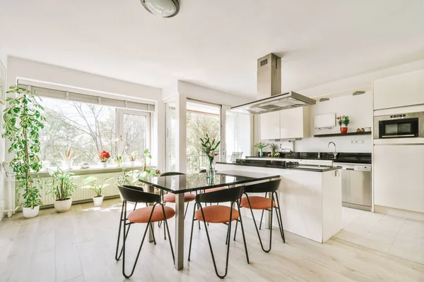 Modern minimalist style interior design of studio apartment with open white kitchen and dining zone with table and chairs illuminated by loft style lamp