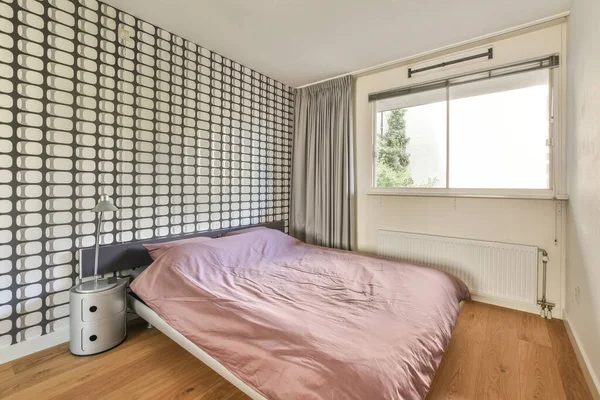 Sunlit bed covered with blanket in light contemporary bedroom with shelves and decorations