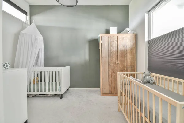 Interior of white room with baby crib and armchair near window with blue chest of drawers in daylight