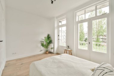 Beyaz çarşaflı rahat bir yatak ve lambasıyla pencere ve balkon kapısının yakınına yerleştirilmiş aydınlık yatak odası.