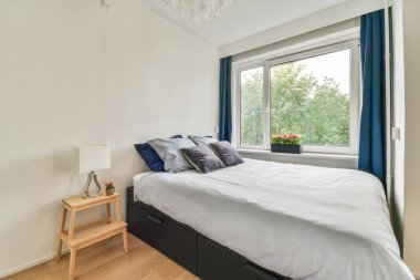 Comfortable bed with white bedclothes and washing machine near window in light bedroom at home