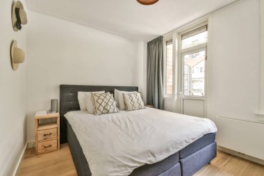 Comfortable bed with white bedclothes and wooden bedside table placed near window in light bedroom at home