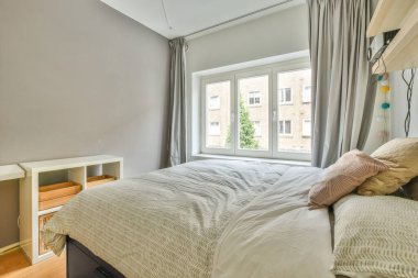 Comfortable bed with white bedclothes and bedside table placed near window in light bedroom at home