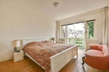 Yatak örtüleriyle rahat bir yatak, yatağın yanında lamba, pencere ve balkon kapısının yanına yerleştirilmiş bir yatak odası.