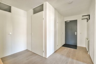 Beyaz duvarları ve kapıları olan küçük koridor modern dairede pencereleri ve parke zemini olan geniş odaya açılıyor.