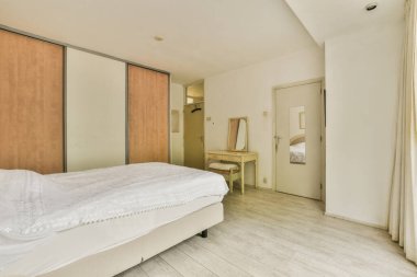 Battaniyeli rahat yatak ve yastıklar küçük yatak odasındaki pencereye dayanan lambayla dolap ve komodinin yanında.