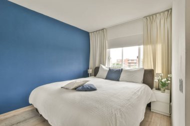 Comfortable bed with white bedclothes and bedside table with lamp placed near window in light bedroom at home