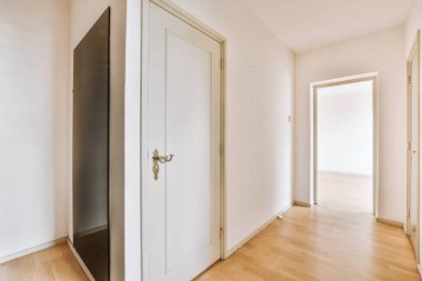 Beyaz duvarları ve kapıları olan dar koridor modern dairede pencereleri ve parke zemini olan geniş odaya açılıyor.