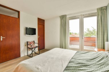 Beyaz çarşaflı rahat bir yatak ve lambasıyla pencere ve balkon kapısının yakınına yerleştirilmiş aydınlık yatak odası.