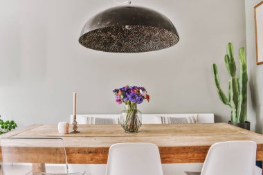 Modern oturma odasındaki siyah kolye lambasının altında çiçek vazosu olan ahşap masa.