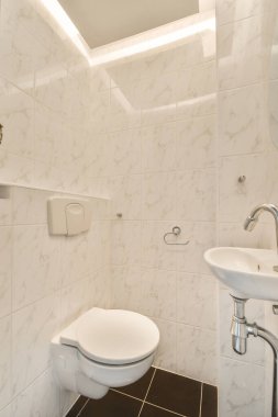 Duvara asılmış tuvalet ve köşedeki bej fayanslı lavabo.