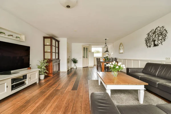 Wooden table with chairs and vase located near comfortable couch against light walls with decorations and TV in modern apartment