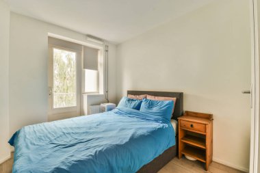 Beyaz çarşaflı rahat bir yatak ve lambasıyla pencere ve balkon kapısının yakınına yerleştirilmiş aydınlık yatak odası.