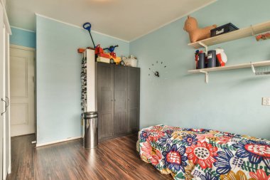 Küçük yatak odasının minimalist iç tasarımı. Köşesinde yumuşak yatak, ahşap gardırop ve lamba aydınlatmalı raflar var.