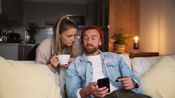 Feliz pareja caucásica sentado en el sofá haciendo pago con tarjeta de crédito en línea en el dispositivo celular — Vídeos de Stock
