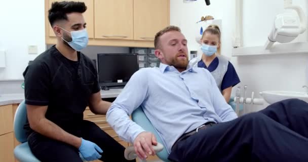 Καυκάσιος άνδρας ασθενής κάθεται στην καρέκλα του γιατρού ενώ άνδρες και γυναίκες νοσηλευτές εξετάζουν τα δόντια — Αρχείο Βίντεο