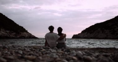 Gün batımında kumsalda kollarında oturan mutlu beyaz çift.