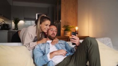 Kanepede oturan mutlu beyaz çift hücresel cihazla selfie çekiyor. 