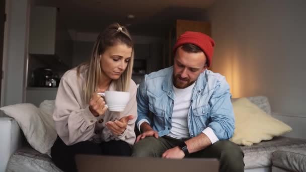 Feliz pareja caucásica sentado en la sala de estar beber café charlando finanzas mientras escribe en el ordenador portátil — Vídeos de Stock