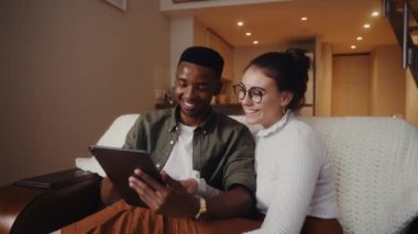 Afrikalı Amerikalı erkek, beyaz kız arkadaşıyla oturmuş dijital tablette resimlere bakarken gülüyor. 