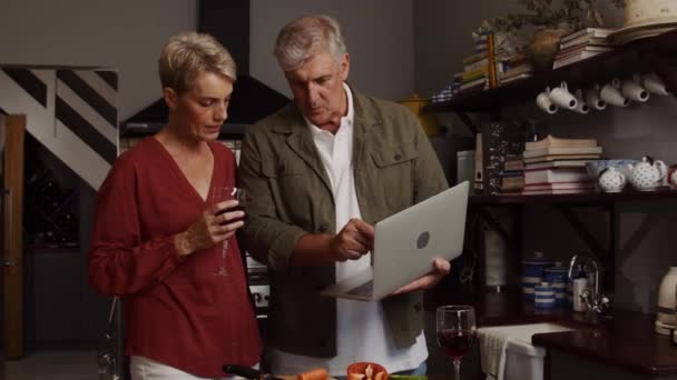 キッチンでコーカシアの高齢者のカップル料理ノートパソコンでレシピを検索しながらロマンチックなディナーを飲むワインを楽しむ — ストック動画